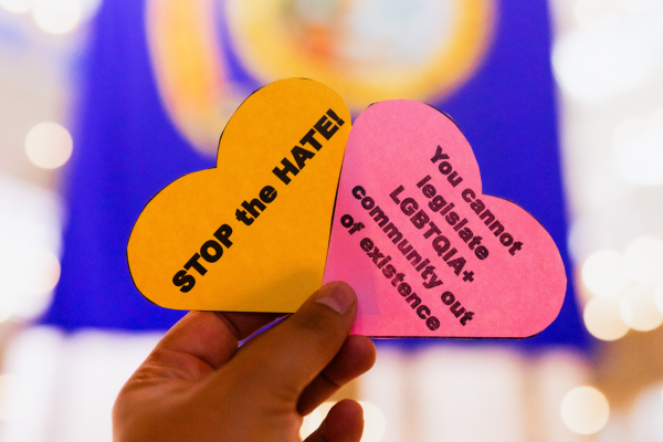 Person holding two hearts with messages that say "stop the hate" and "you cannot legislate LGBTQIA+ community out of existence." The State of Idaho flag is blurred in the background at the Idaho Capitol.