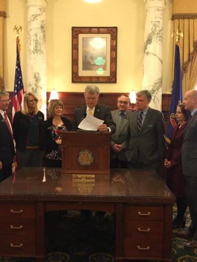 Gov. Otter standing at a podium signing a bill into law in the Governor's office with people standing around him