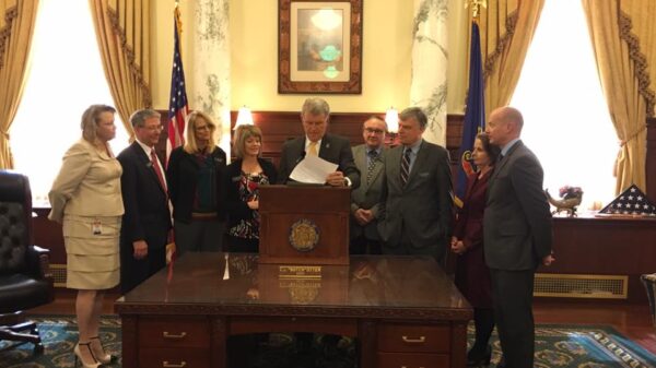 Gov. Otter standing at a podium signing a bill into law in the Governor's office with people standing around him