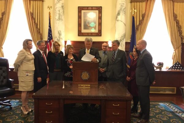 Gov. Otter standing at a podium signing a bill into law in the Governor's office with people standing around him