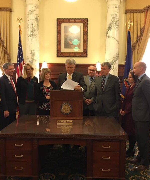 Gov. Otter standing at a podium signing a bill into law in the Governor's office with people standing around him