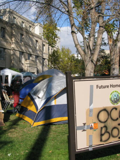 a sign with a cardboard sign taped on top with spray painted "occupy boise" on it with tents in the background