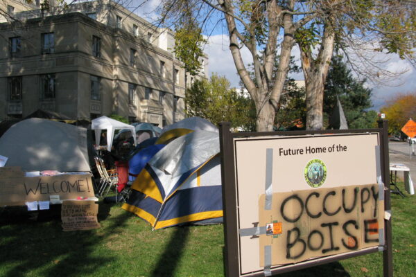 a sign with a cardboard sign taped on top with spray painted "occupy boise" on it with tents in the background