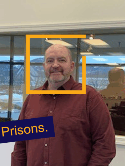 John is a middle-aged balding white man looking at the camera and smiling from in front of a business's wall and window into an office.