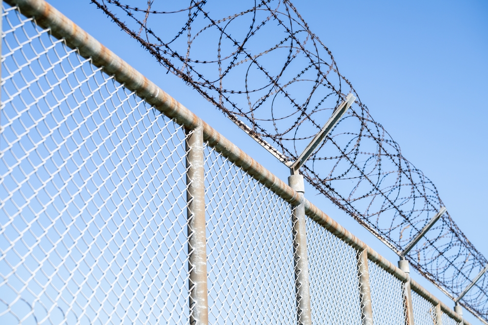 Large fence with barb wire at the top, often seen surrounding prisons.