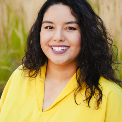 Rosseli is a Latine woman with black hair wearing a yellow shirt with a green and brown nature background.