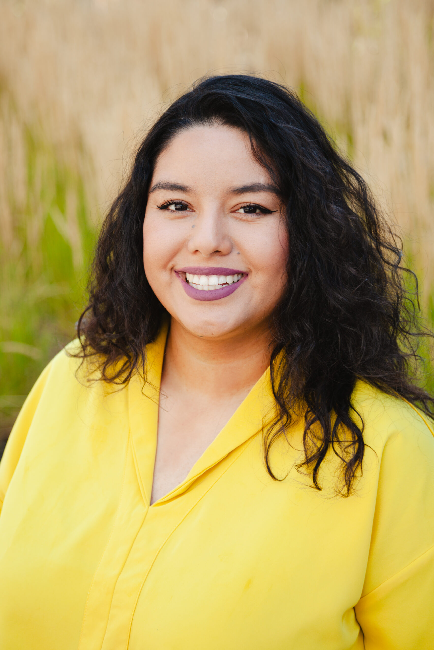 Rosseli is a Latine woman with black hair wearing a yellow shirt with a green and brown nature background.