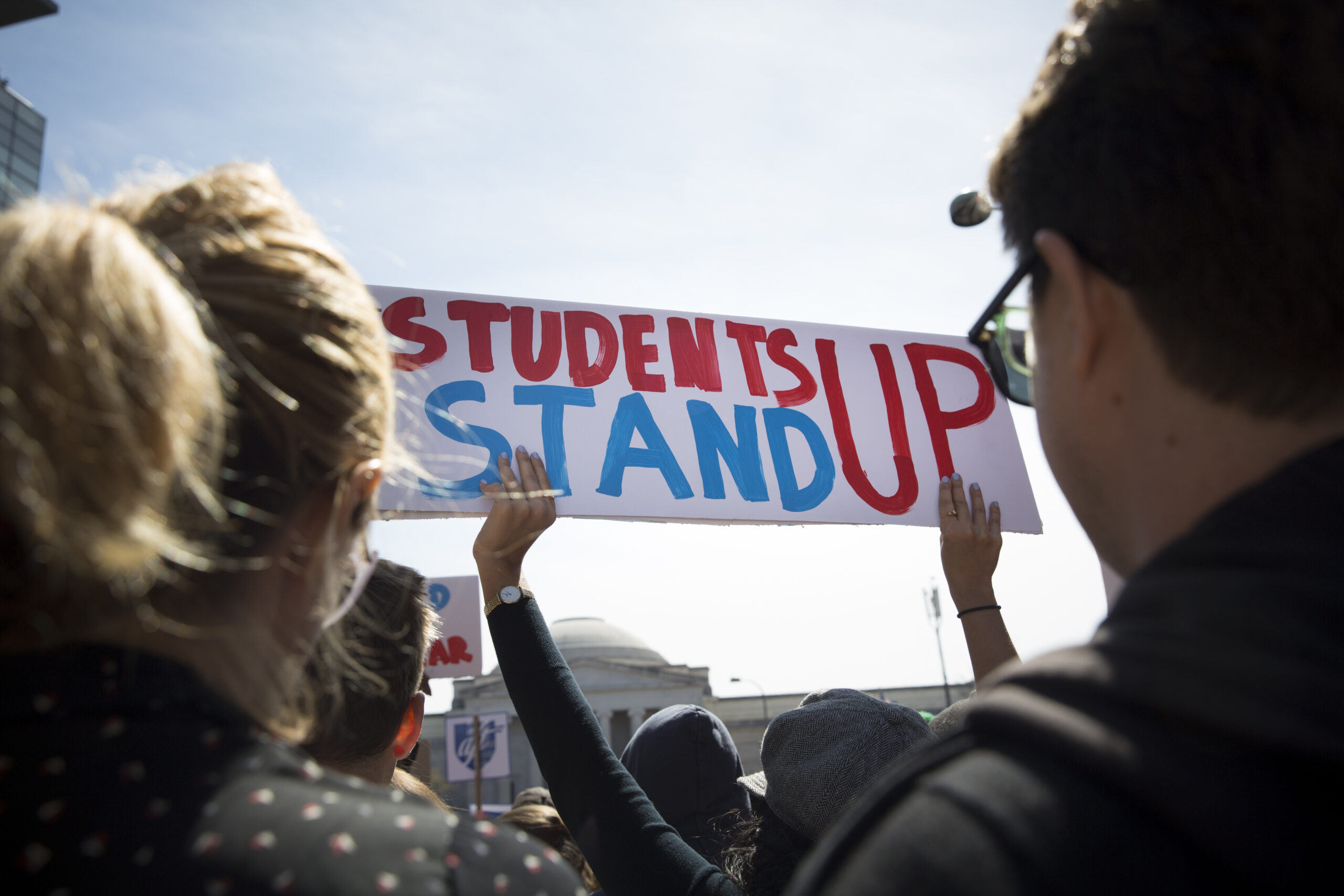 a person is holding up a white banner that reads "students stand up" in read and blue letters. In the foreground there are two people with their backs to us to either side of the sign.