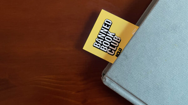 The top left corner of a blue book sits on a brown surface. A Yellow bookmark stuck our of the top of the book with the words "Banned Books Club ACLU" in white and black letters.