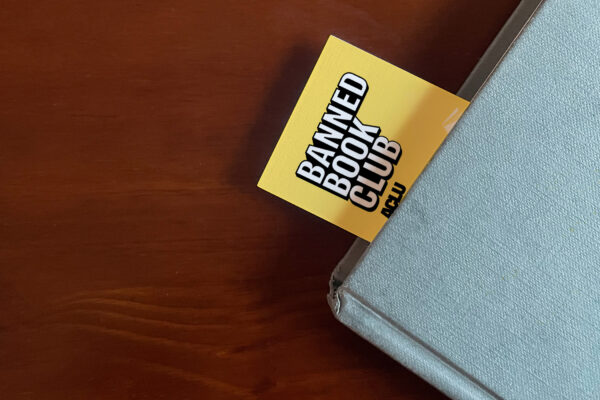 The top left corner of a blue book sits on a brown surface. A Yellow bookmark stuck our of the top of the book with the words "Banned Books Club ACLU" in white and black letters.