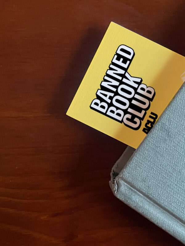 The top left corner of a blue book sits on a brown surface. A Yellow bookmark stuck our of the top of the book with the words "Banned Books Club ACLU" in white and black letters.