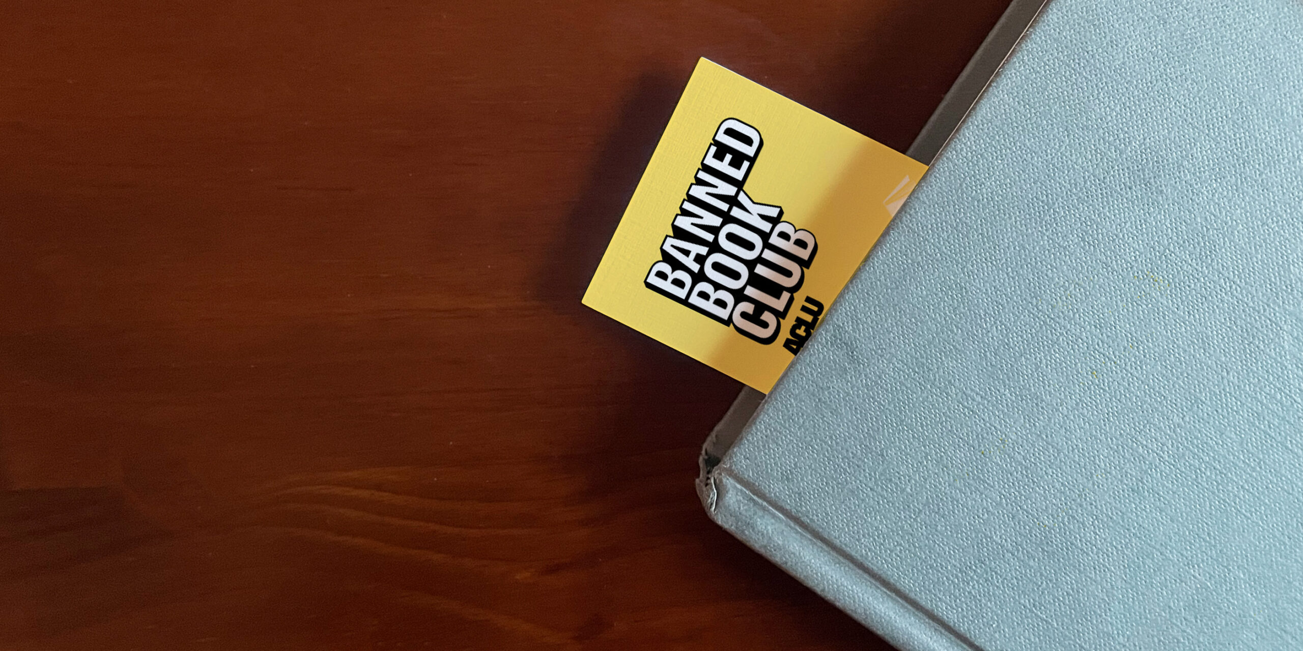 The top left corner of a blue book sits on a brown surface. A Yellow bookmark stuck our of the top of the book with the words "Banned Books Club ACLU" in white and black letters.