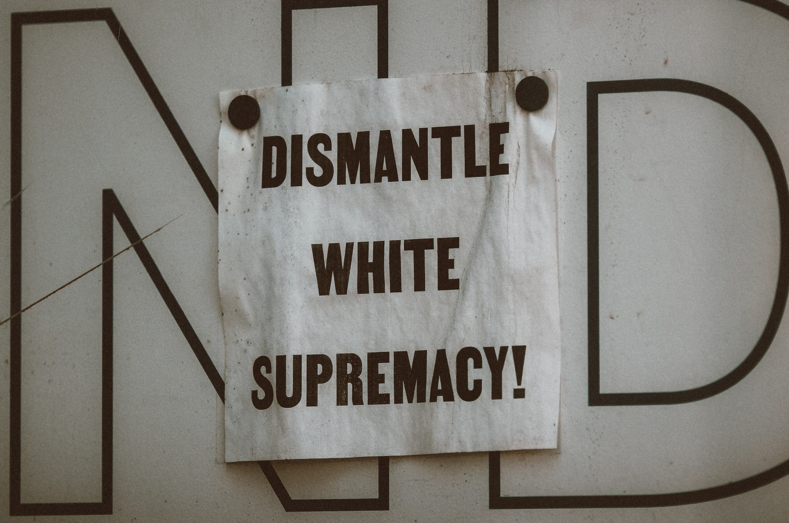 A white sign with the words "dismantle white supremacy" in black letters is tacked to a white wall wall in front of the letters n and d.