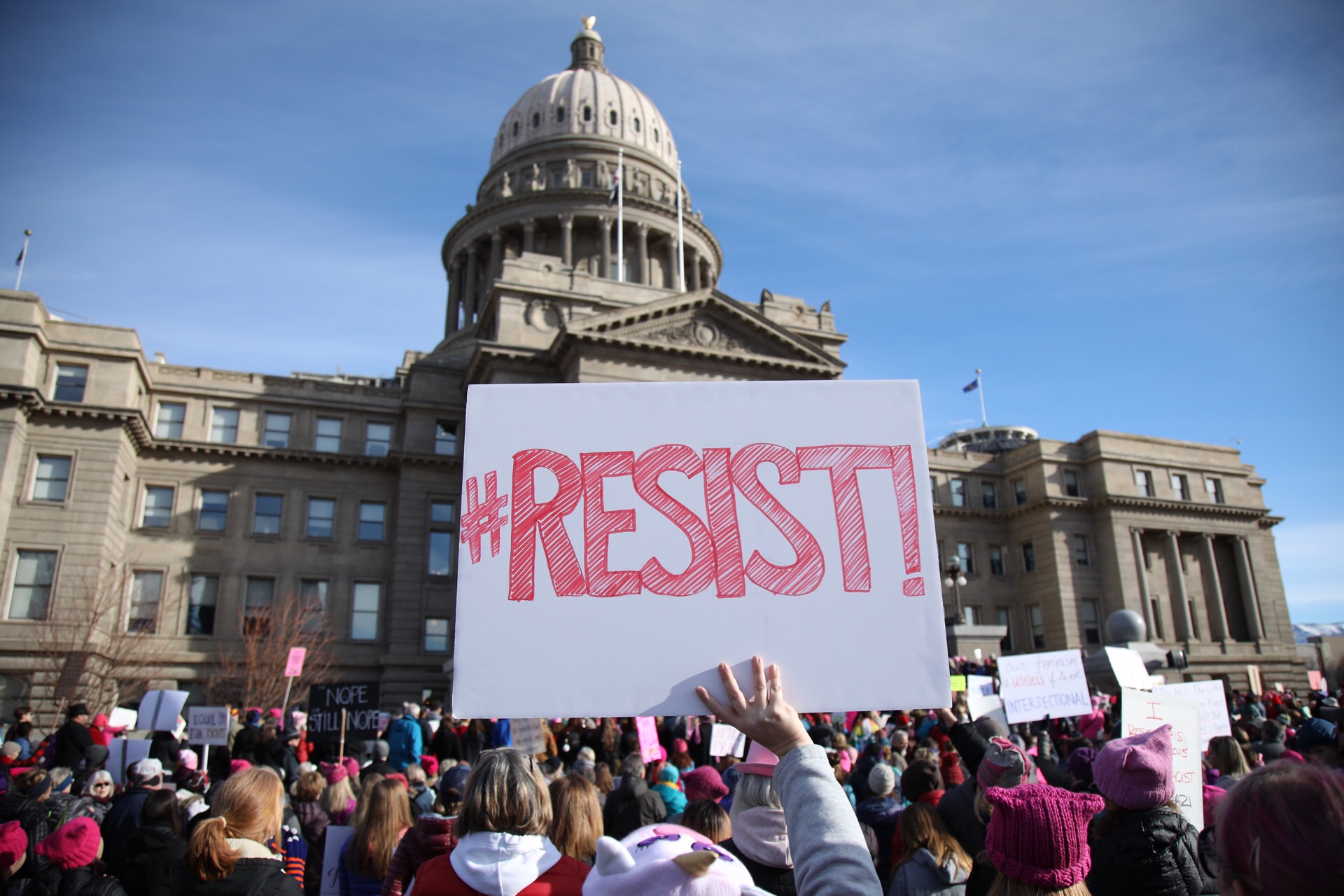 In the center of the image, a person is holding a white sign with "#Resists" in red letters there is a crowd of people in front  of the Idaho state capitol building in the background