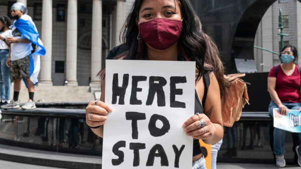 A young latine girl wears a mask and holds a sign that reads "here to stay"