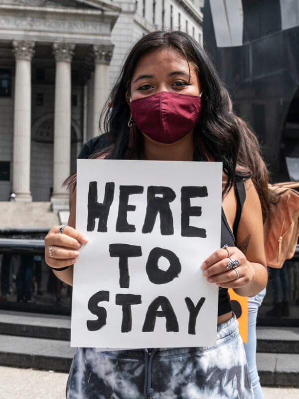 A young latine girl wears a mask and holds a sign that reads "here to stay"
