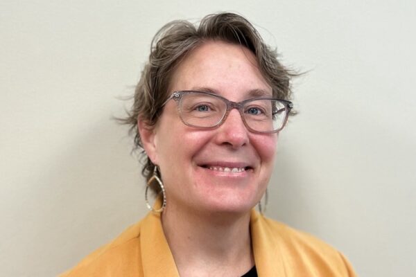 A phot of Julianne wearing grey framed glasses, round earrings in a yellow blazer with a black shirt. Julianne is smiling.