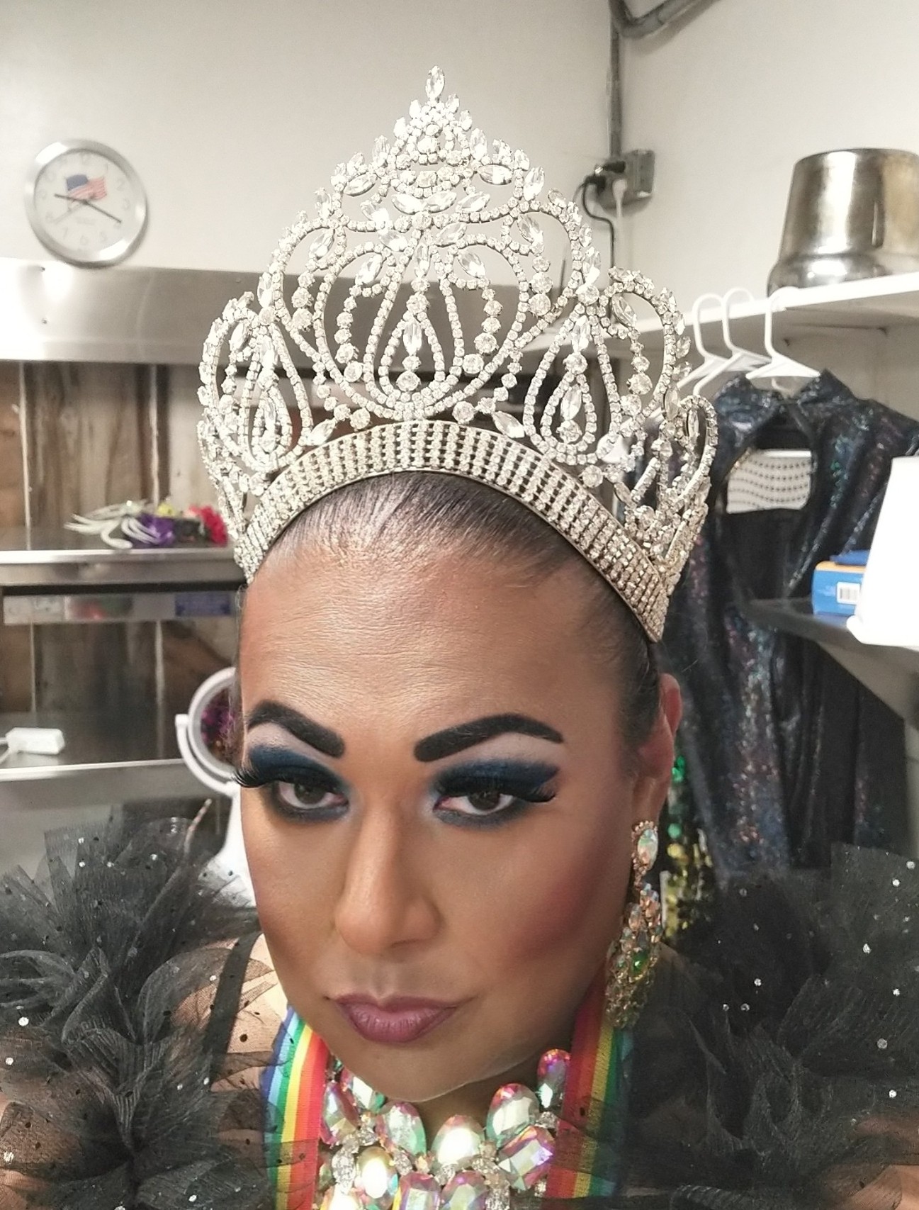 A photo of Crystlll Blue from the shoulders up. She is looking directly at the camera and is wearing a black ruffled top with colorful necklaces. She is wearing a tiara with bright rhinestones.