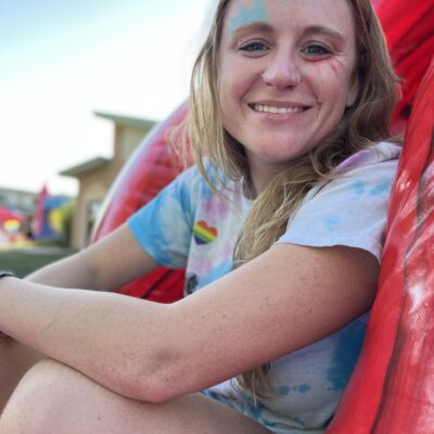 A picture of Jenna sitting down with their arms resting on their knees. Jenna is smiling and wearing a tie-dye t-shirt. Jenna has colorful face paint above their left eye and below their right cheek.