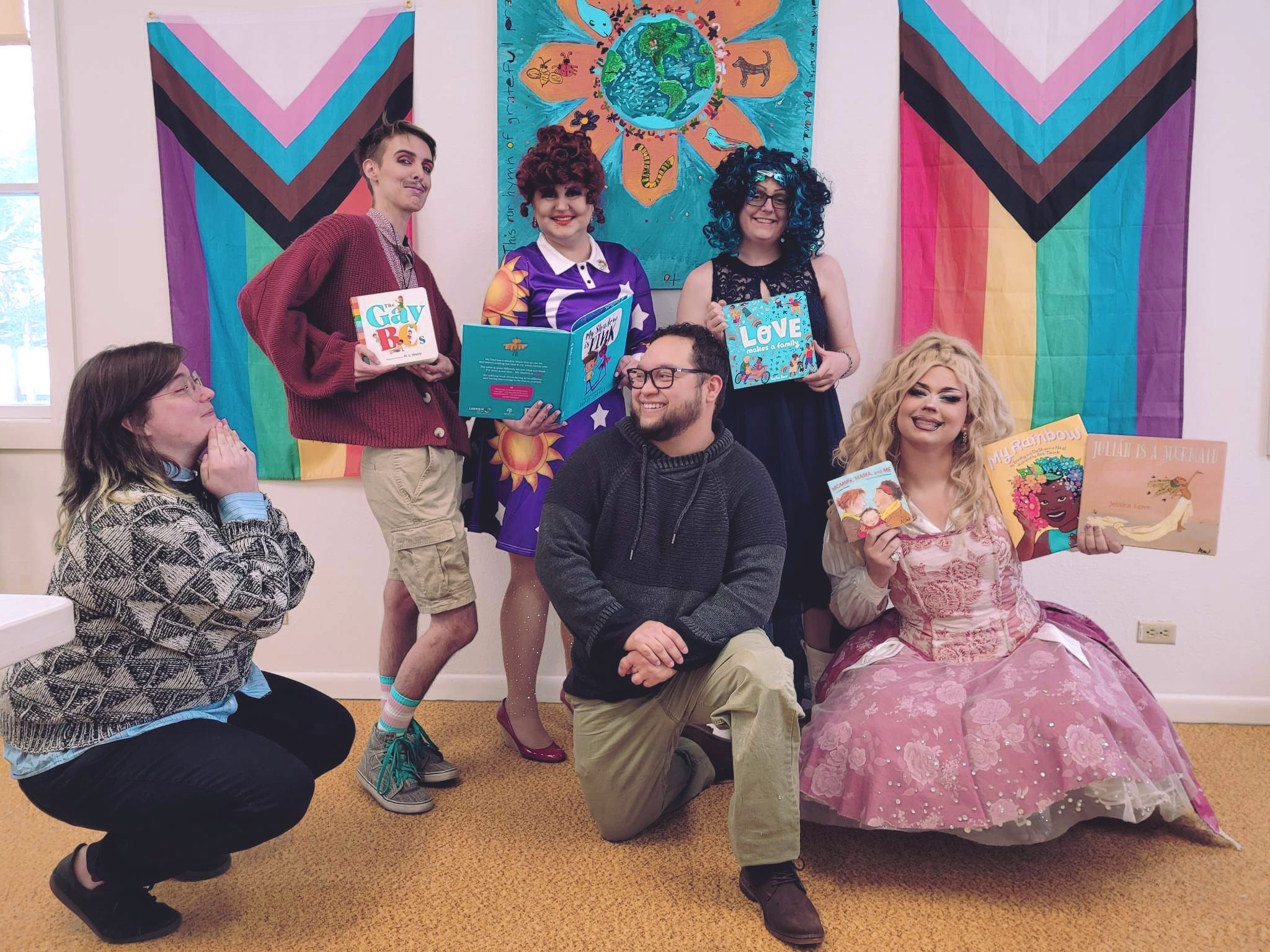 A group of 6 drag performers in various poses holding books in front of two progress pride flags and a colorful banner with the sun.