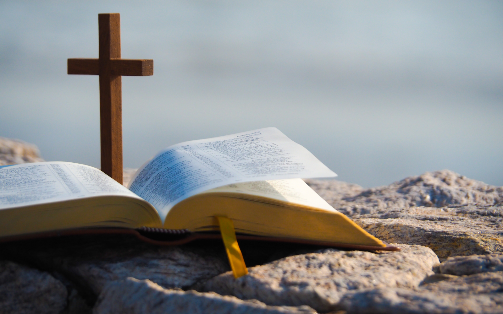 A wooden cross stands up on top of a Bible, both sitting on a rock with a blurred background