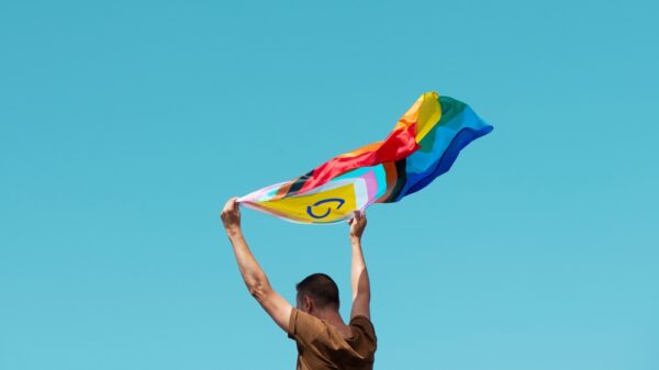 White man hold a pride flag up in the air with a blue sky in the background