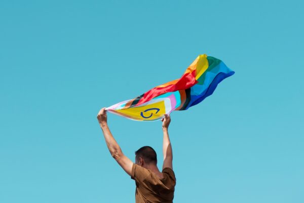 White man hold a pride flag up in the air with a blue sky in the background