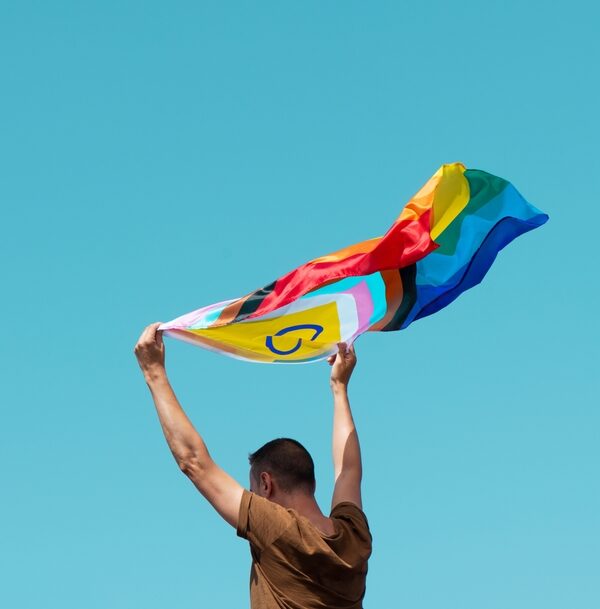 White man hold a pride flag up in the air with a blue sky in the background
