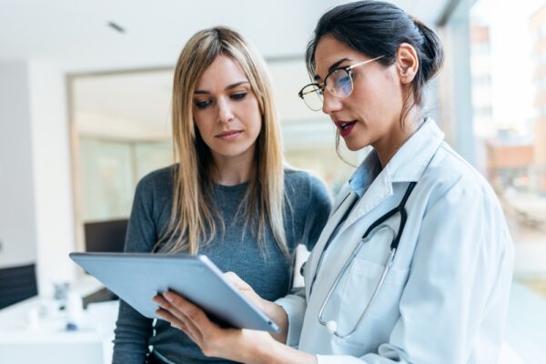 Female doctor is explaining something to a female patient while looking at a clipboard.