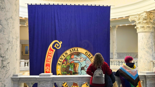 Two people stand in the Idaho Capitol looking at the State of Idaho flag, with one person wearing a Pride flag