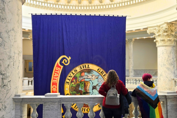 Two people stand in the Idaho Capitol looking at the State of Idaho flag, with one person wearing a Pride flag