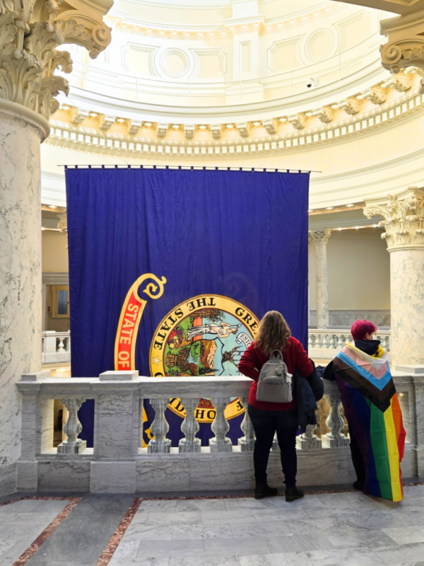 Two people stand in the Idaho Capitol looking at the State of Idaho flag, with one person wearing a Pride flag