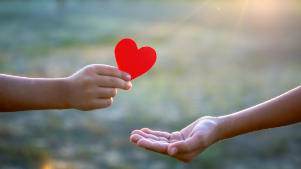 Person handing a heart to another hand with green, grassy background