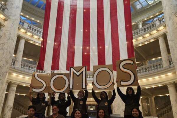 Picture of American flag inside the rotunda of the Idaho Capitol with Black and Brown people at the bottom wearing brown SOMOS shirts and holding signs that spell out SOMOS IDAHO.