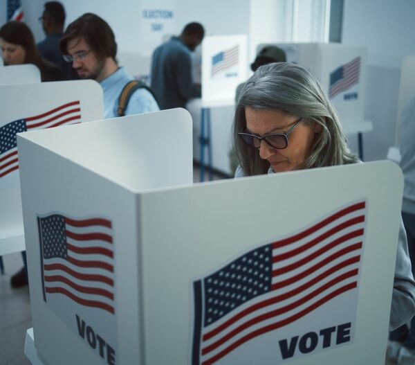 Multiple people of various ages voting in ballot boxes that have an American flag and the word "vote" on them