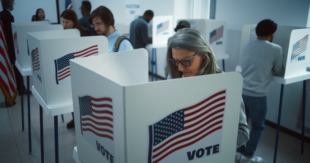 Multiple people of various ages voting in ballot boxes that have an American flag and the word "vote" on them