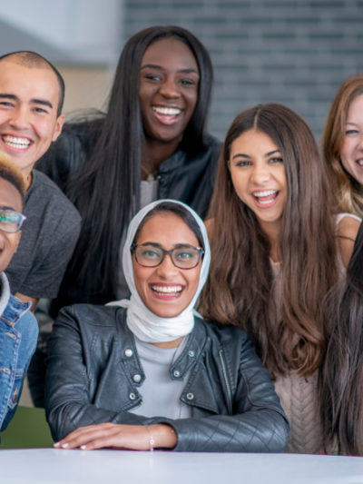 A group of seven individuals looks at the camera smiling. They are diverse in gender, race, religion, and ethnicity with men and women representing Black, Indigenous, Asian, and white races, and one woman wears a hijab.