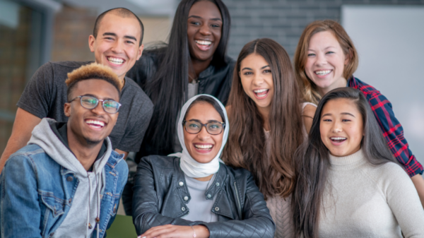 A group of seven individuals looks at the camera smiling. They are diverse in gender, race, religion, and ethnicity with men and women representing Black, Indigenous, Asian, and white races, and one woman wears a hijab.