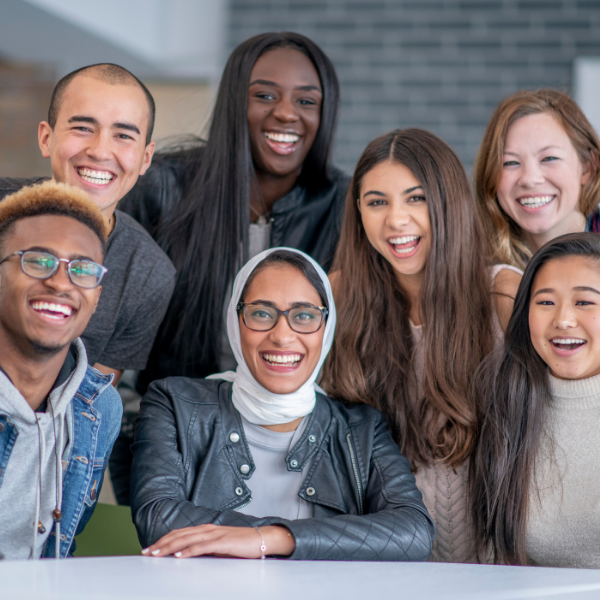 A group of seven individuals looks at the camera smiling. They are diverse in gender, race, religion, and ethnicity with men and women representing Black, Indigenous, Asian, and white races, and one woman wears a hijab.