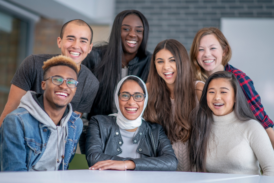 A group of seven individuals looks at the camera smiling. They are diverse in gender, race, religion, and ethnicity with men and women representing Black, Indigenous, Asian, and white races, and one woman wears a hijab.