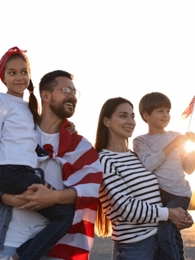 A man holding a little girl stands next to a woman holding a little boy with the USA flag draped over them, looking happy together.