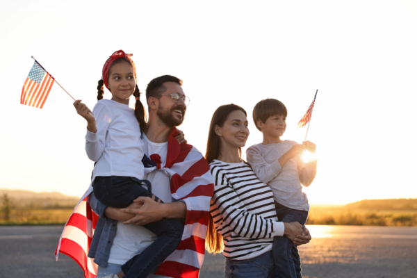 A man holding a little girl stands next to a woman holding a little boy with the USA flag draped over them, looking happy together.