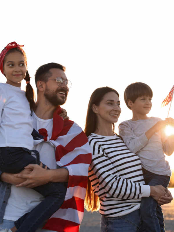 A man holding a little girl stands next to a woman holding a little boy with the USA flag draped over them, looking happy together.