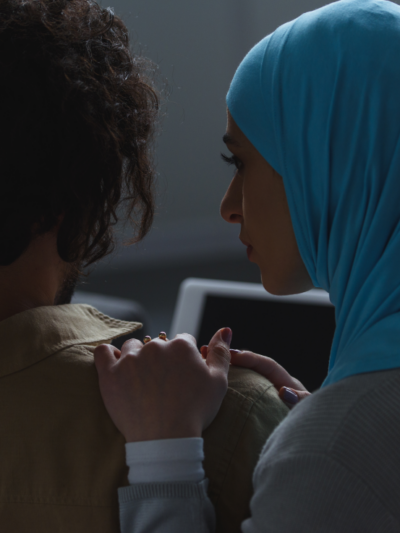 A woman wearing a hijab comforts at a man with shoulder-length hair who is facing away from the camera.