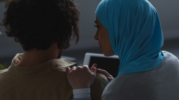 A woman wearing a hijab comforts at a man with shoulder-length hair who is facing away from the camera.