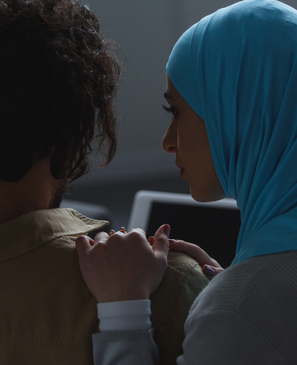 A woman wearing a hijab comforts at a man with shoulder-length hair who is facing away from the camera.