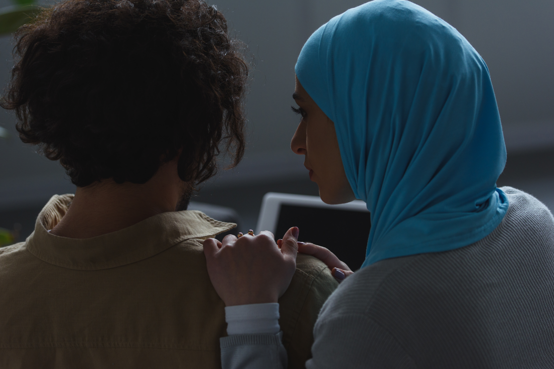 A woman wearing a hijab comforts at a man with shoulder-length hair who is facing away from the camera.