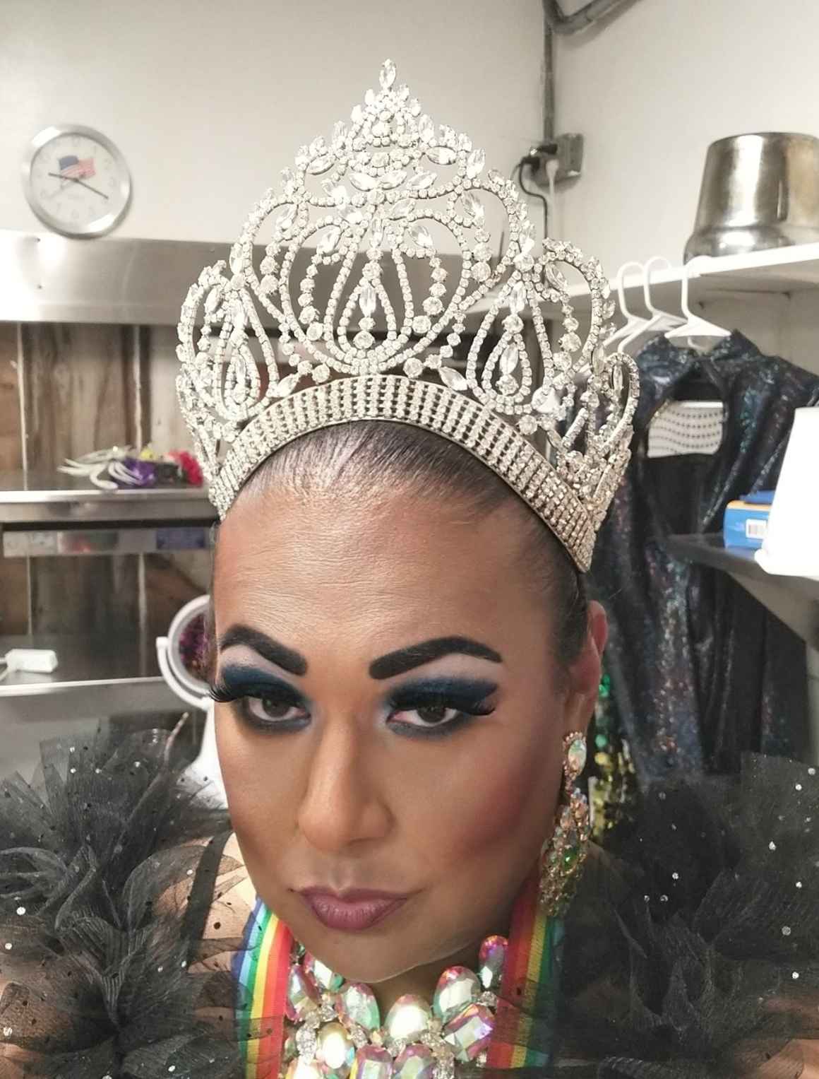 A photo of Crystlll Blue from the shoulders up. She is looking directly at the camera and is wearing a black ruffled top with colorful necklaces. She is wearing a tiara with bright rhinestones. 