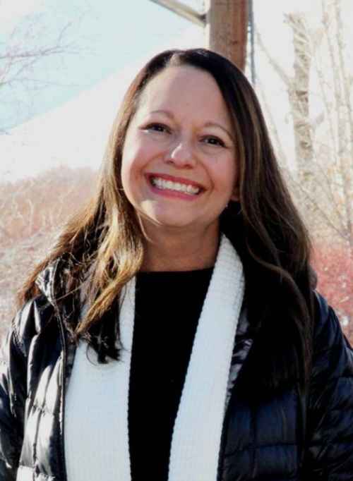 A photo of Deborah smiling. She is wearing a black jacket and a white scarf and black shirt. There are trees in the background.