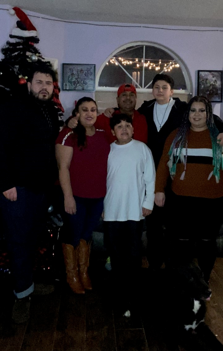 A photo of Elizabeth and her family in portrait mode. Elizabeth is on the right wearing a red shirt with jeans and brown boots. They are in a living room. There is a Christmas tree and Christmas lights in the background.